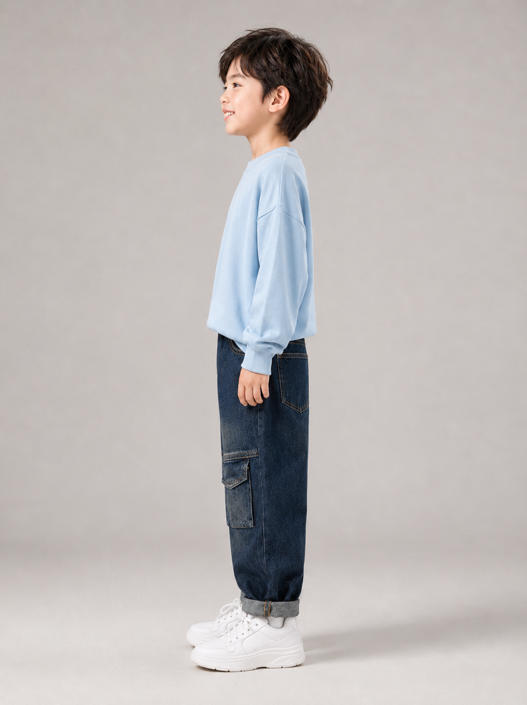 A child model shown in profile wearing a light blue crewneck sweatshirt, dark blue cargo jeans, and white sneakers against a neutral gray studio background.