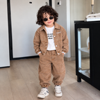 A child wearing a matching brown corduroy jacket and pants set with a white graphic t-shirt and white sneakers, standing in a modern indoor room.